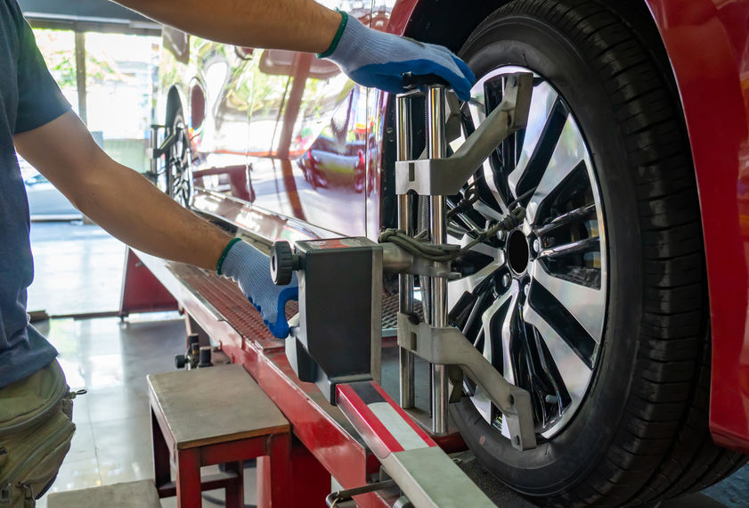 A mechanic adjusts the wheel alignment of a red car using specialized equipment in a garage setting.