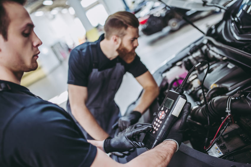 Two mechanics use a diagnostic tool to inspect a car engine in a garage setting.