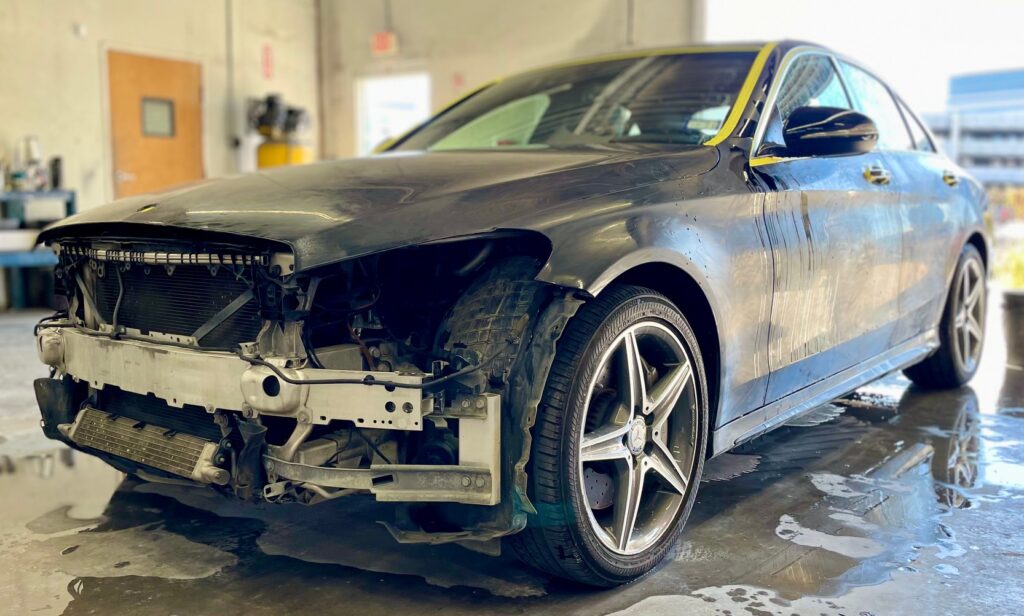 A damaged black sedan without a front bumper is parked in a workshop with its headlights and grille exposed.